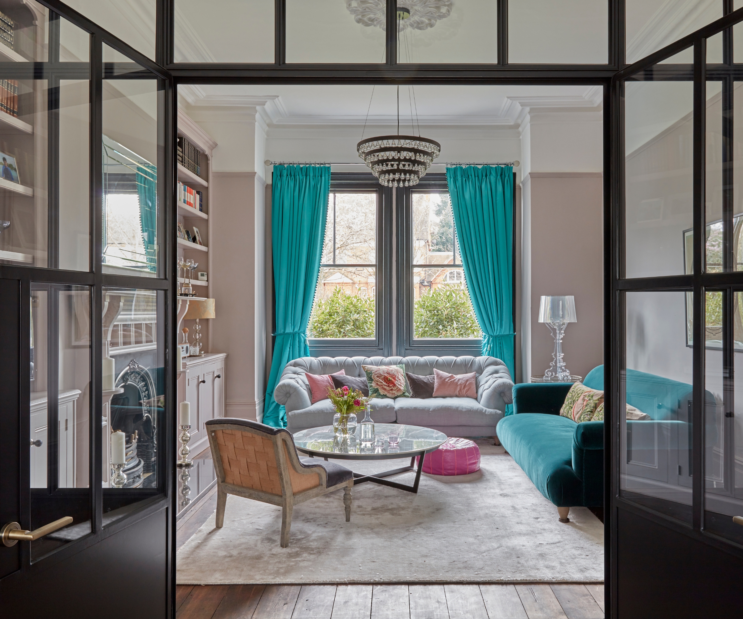 Living room seen through black-framed glass doors, grey and teal velvet sofas, teal curtains, chandelier.