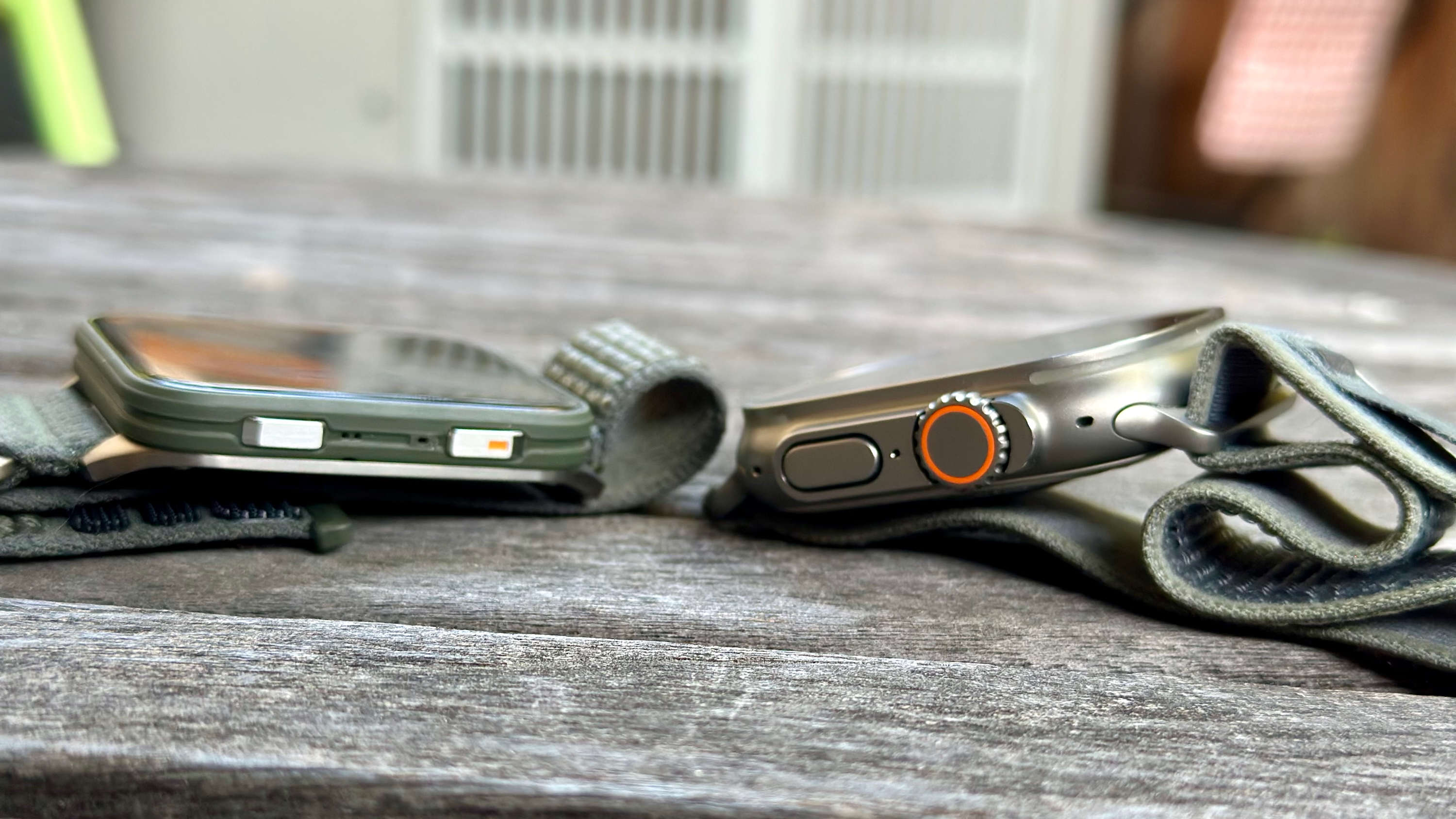 The Garmin Venu X1 (left) and Apple Watch Ultra 2 (right) side by side on a table, showing the relative skinnyness of the X1 compared to the thick Ultra 2.