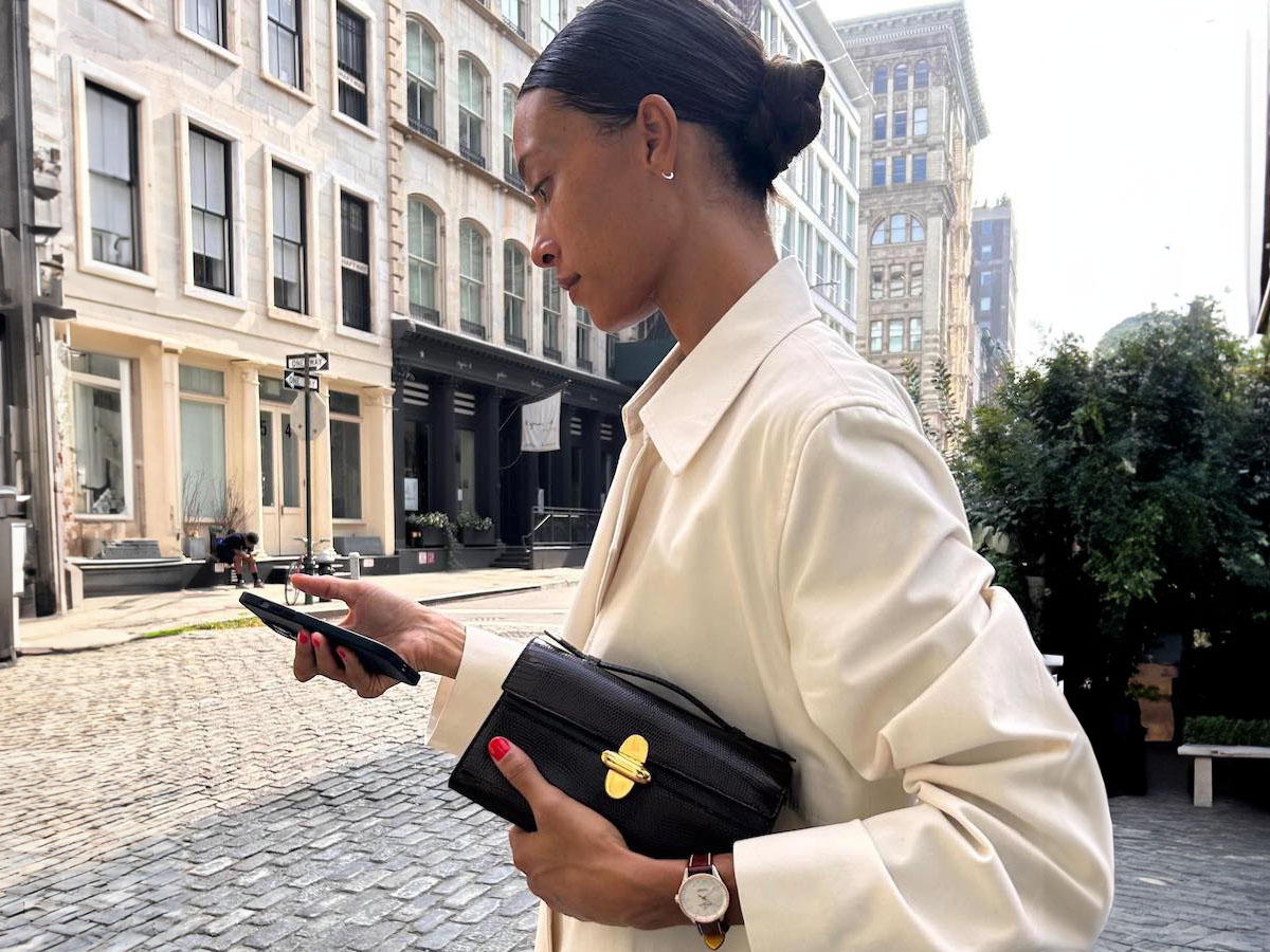 Fashion influencer Tylynn Nguyen poses on the cobblestone streets of Soho in New York City looking at her cellphone wearing a cream jacket, black Savette bag, and Breitling watch.