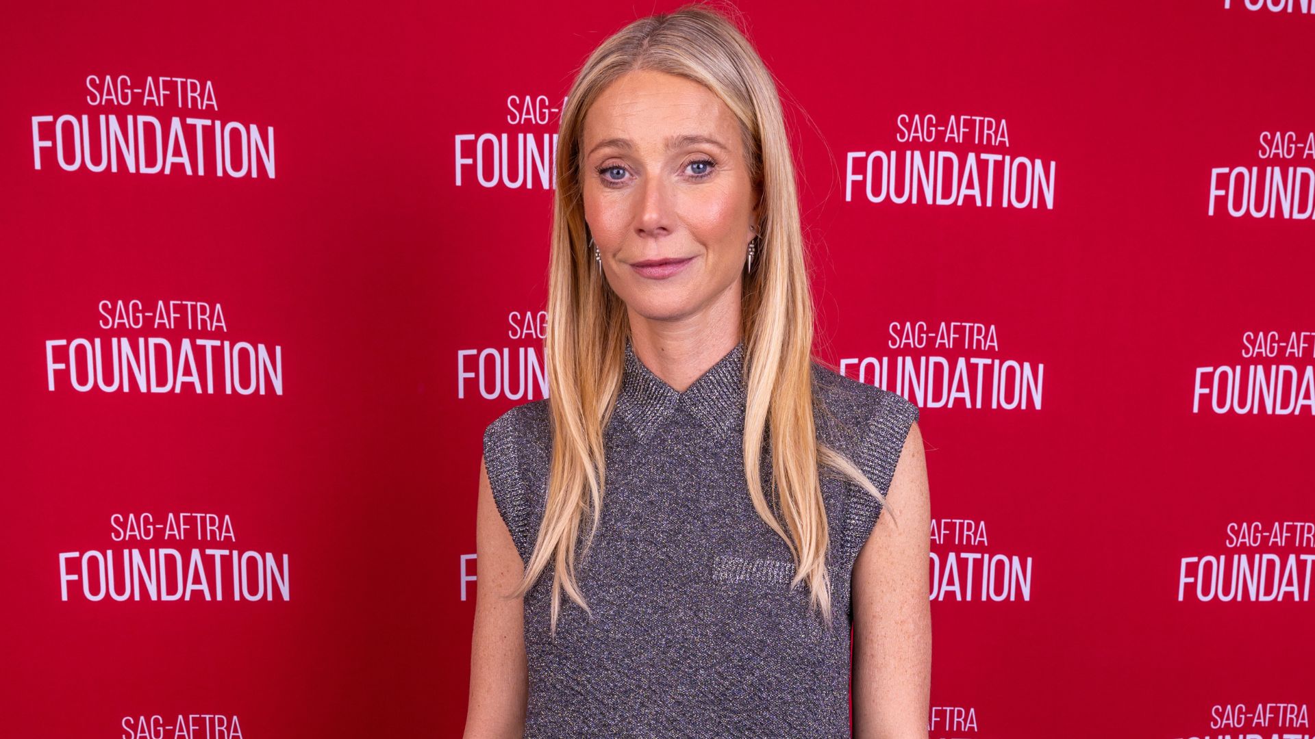 Gwyneth Paltrow poses on a red carpet wearing a grey knitted top with trousers and navy heels.