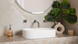 A white oval countertop basin is set on a marble-effect surface with warm beige and cream tones. Above the sink, a large round mirror reflects the soft lighting, complementing the textured wall tiles in a neutral shade. 