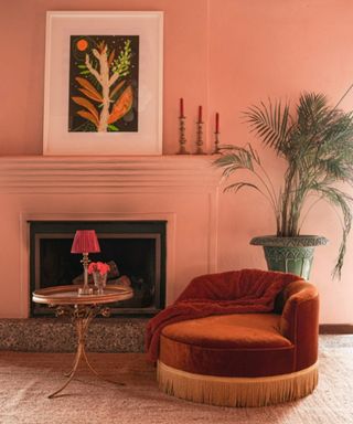 pink wall with fireplace and artwork, large houseplant, orange round chair and ornate side table