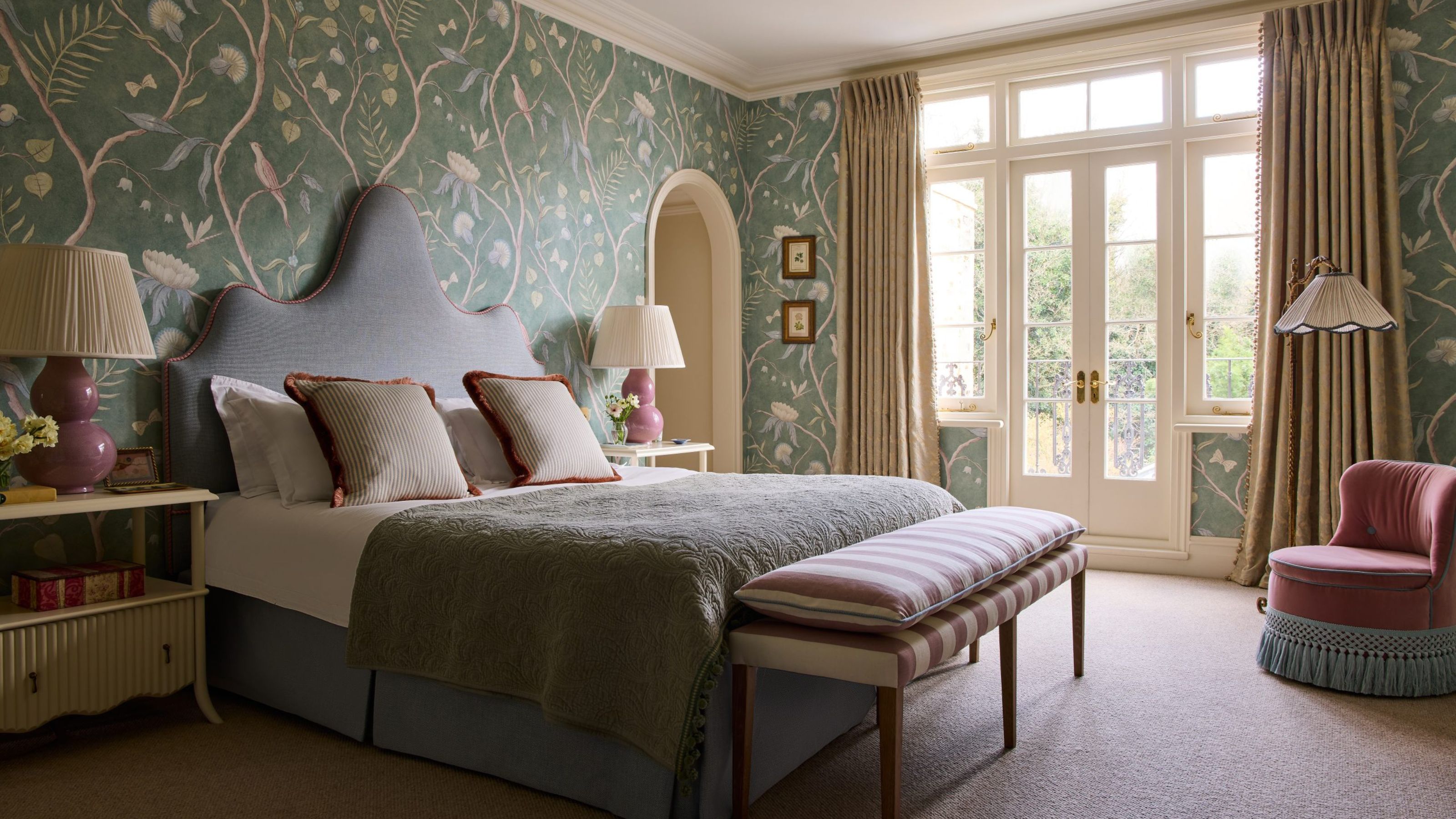 Elegant bedroom with green floral wallpaper, a uniquely shaped grey headboard, a striped bench at the foot of the bed, a pink fringed armchair, and large French doors.
