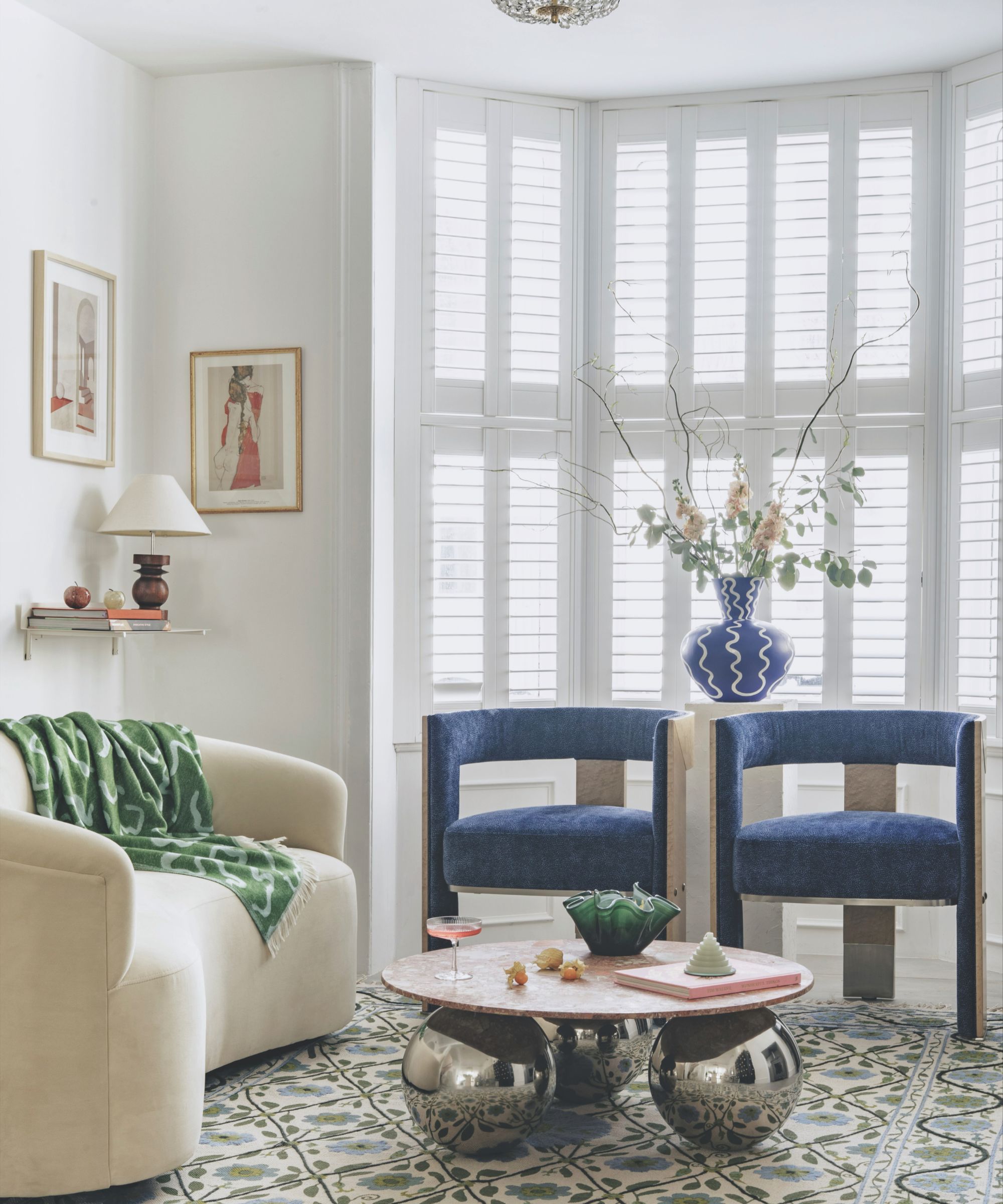A bright living room with a curved cream sofa, blue velvet chairs, and a marble coffee table on a floral rug, set against white shuttered bay windows.