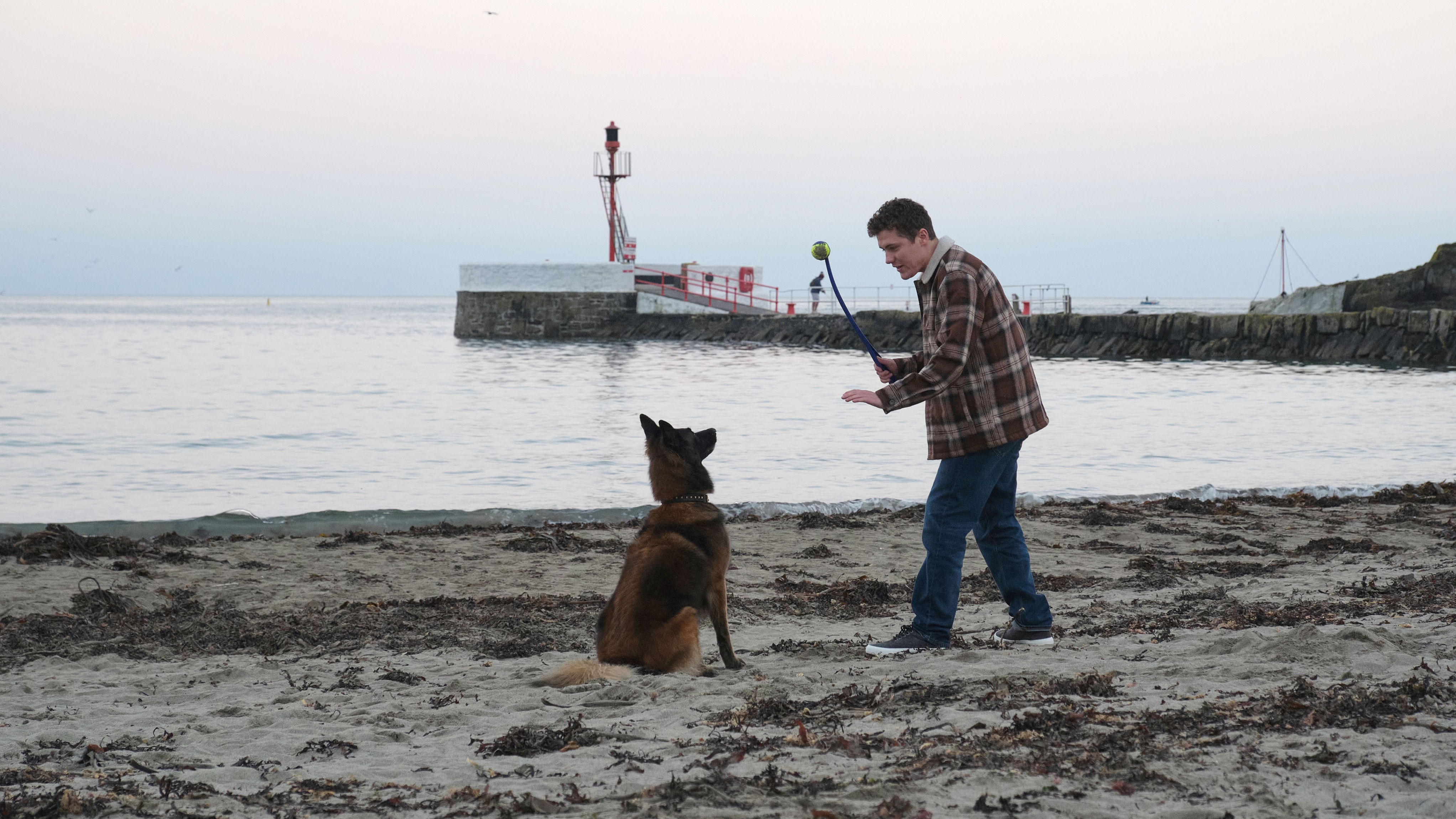 PC Kelby Hartford (Dylan Llewellyn), in his casual clothes of a plaid jacket and blue jeans, is on the beach with a large black and chestnut coloured dog. The dog is sitting obediently and Kelby is waving a stick as if he's about to throw it.