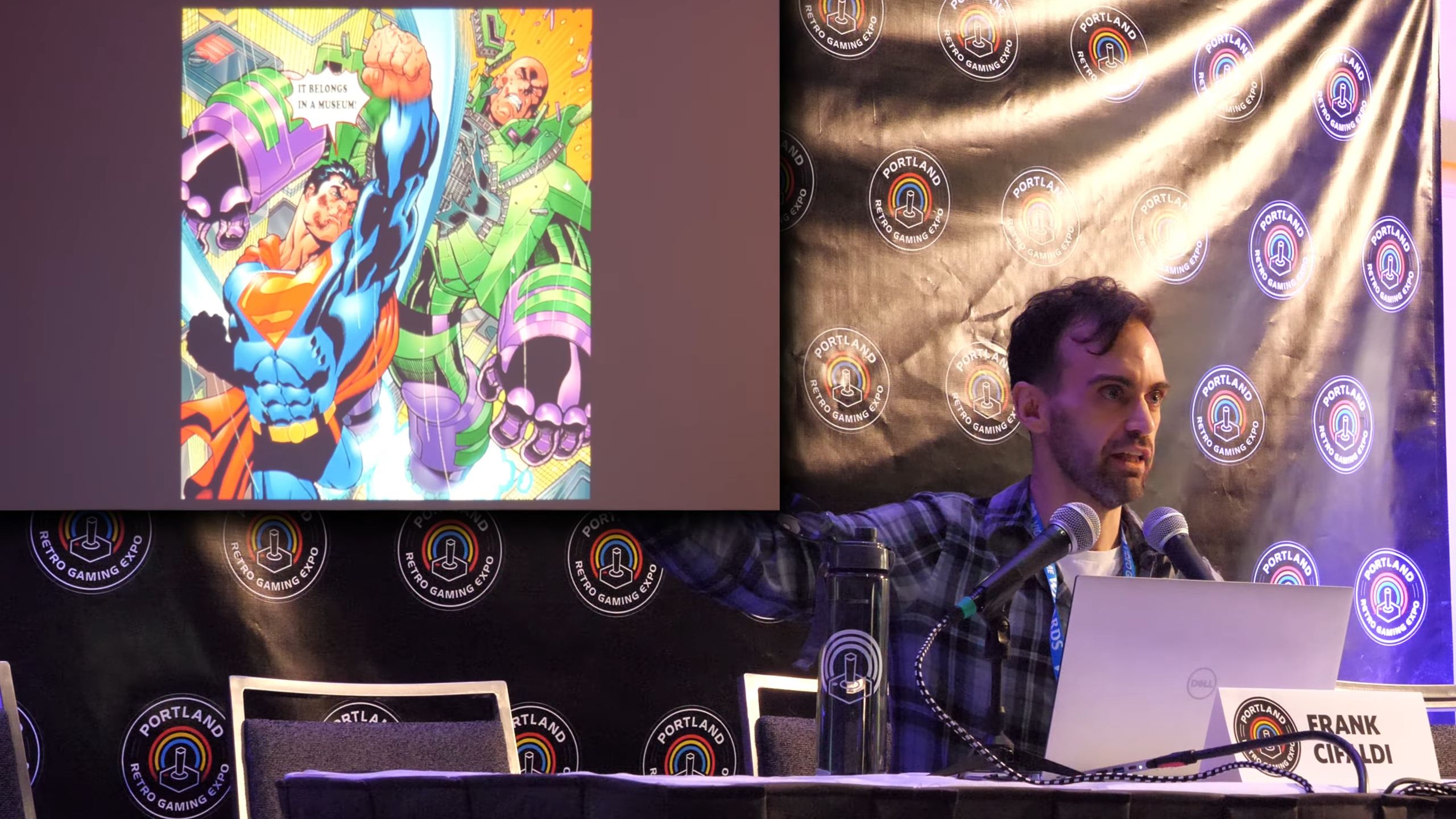 A presentation featuring an illustration of Superman and Lex Luthor on a screen, with Frank Cifaldi speaking at a table in front. The screen shows bright colors and the text, "IT BELONGS IN A MUSEUM!"