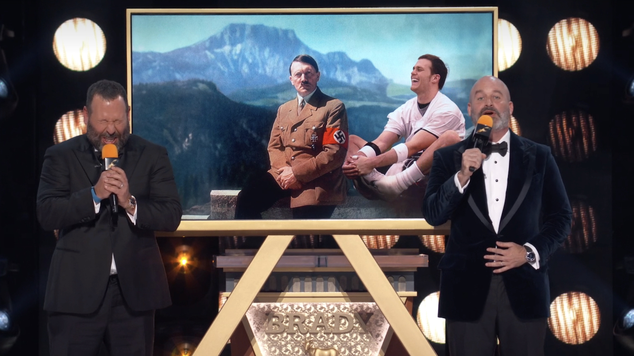 Bert Kreischer laughing next to Tom Segura at The Roast of Tom Brady as they stand in front of a doctored photo of Tom Brady sitting next to Adolf Hitler in front of a mountain.