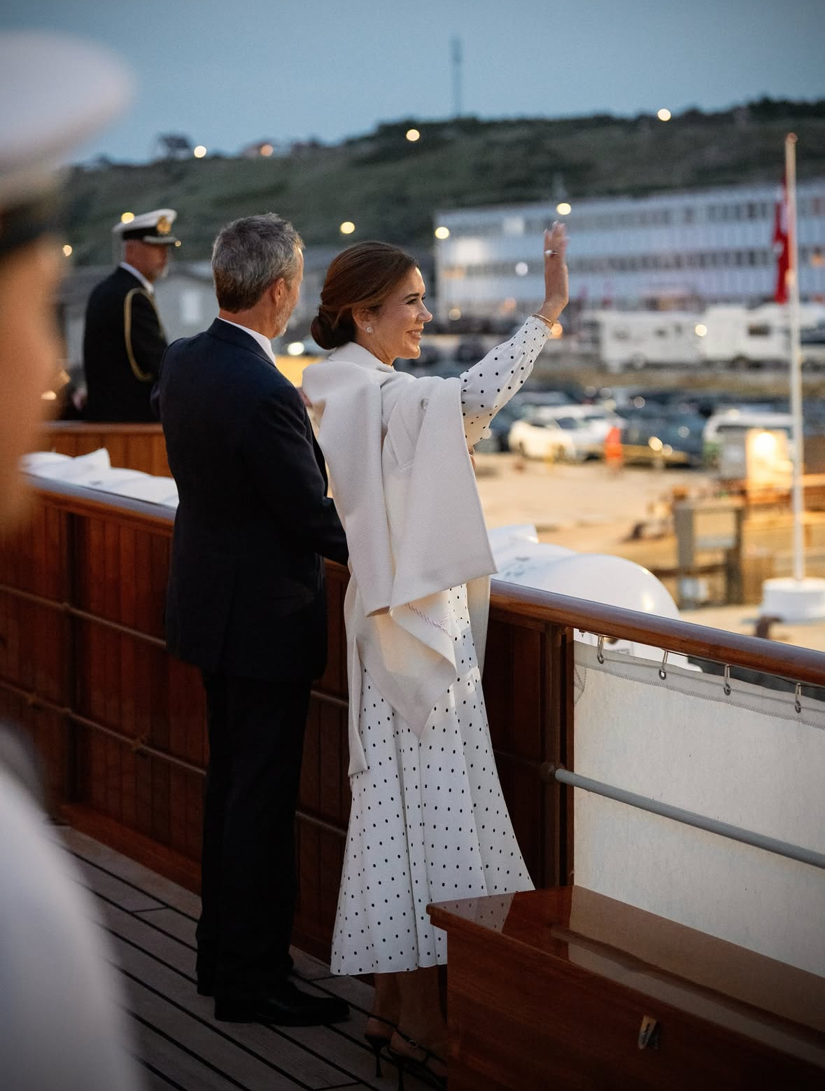 Queen Mary standing next to King Frederik, wearing a white polka dot dress on a ship and waving with her back to the camera