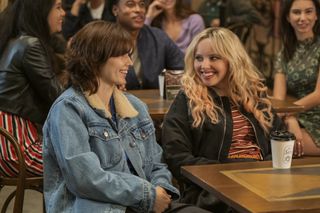 Ruby Cruz as Ash and Mia Rodgers as Taylor, sitting at a table in a crowded café, in 'The Sex Lives of College Girls' season 4.