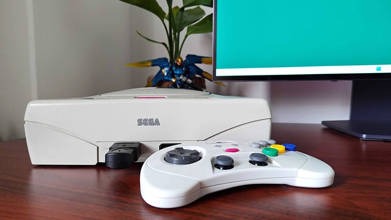 Retro-bit Sega Saturn controller next to White Sega Saturn on woodgrain desk with plant and monitor in backdrop.
