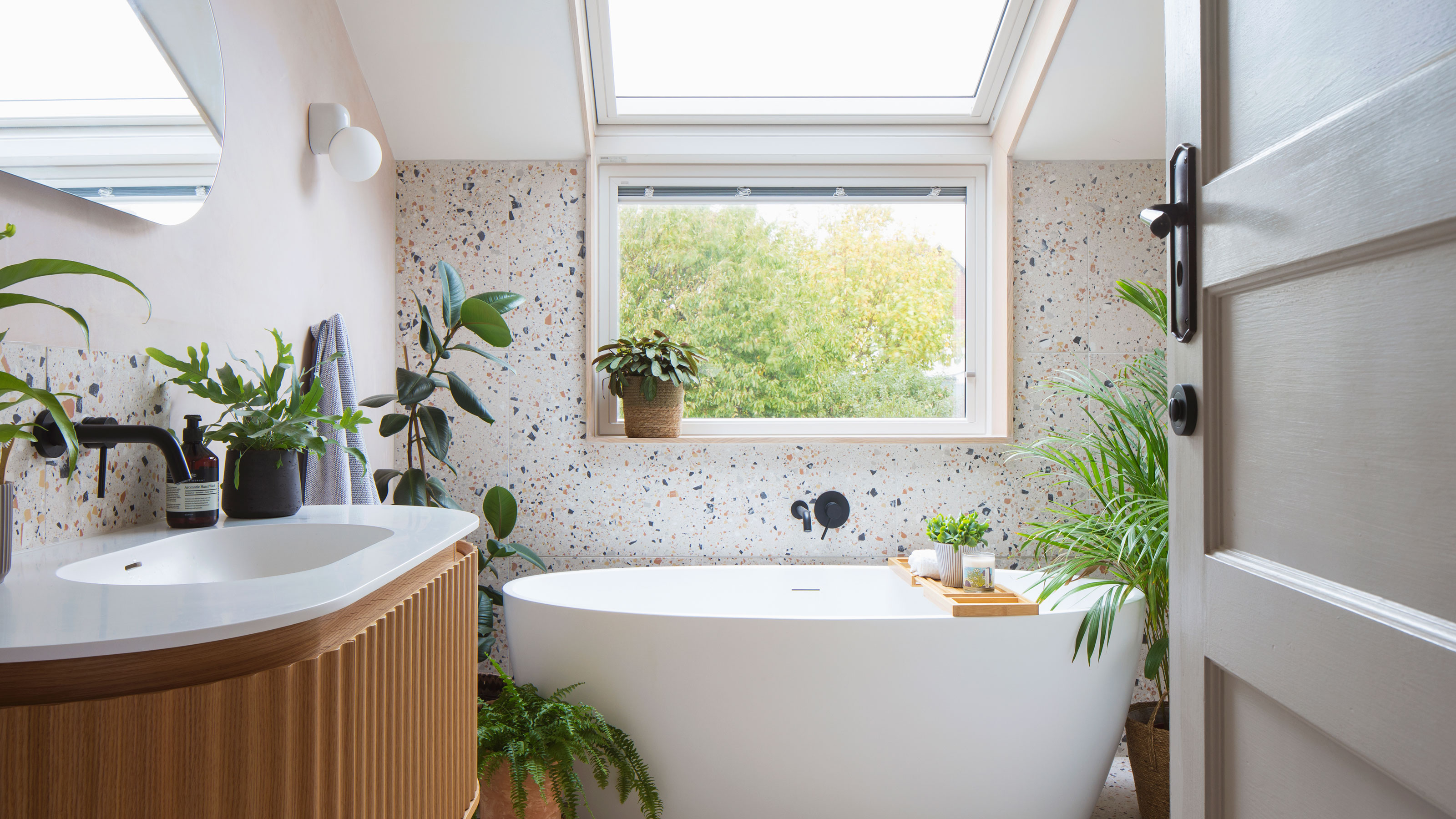 Bathroom with terrazzo on wall and floor, a fluted wooden vanity with white countertop, a freestanding bathtub, pink wall with lozenge shaped mirror and a wall mounted light.