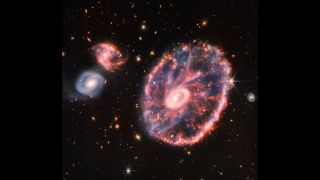 a large galaxy shown shining pink, with two nearby galaxies to the upper left. dots of far distant galaxies are littered about