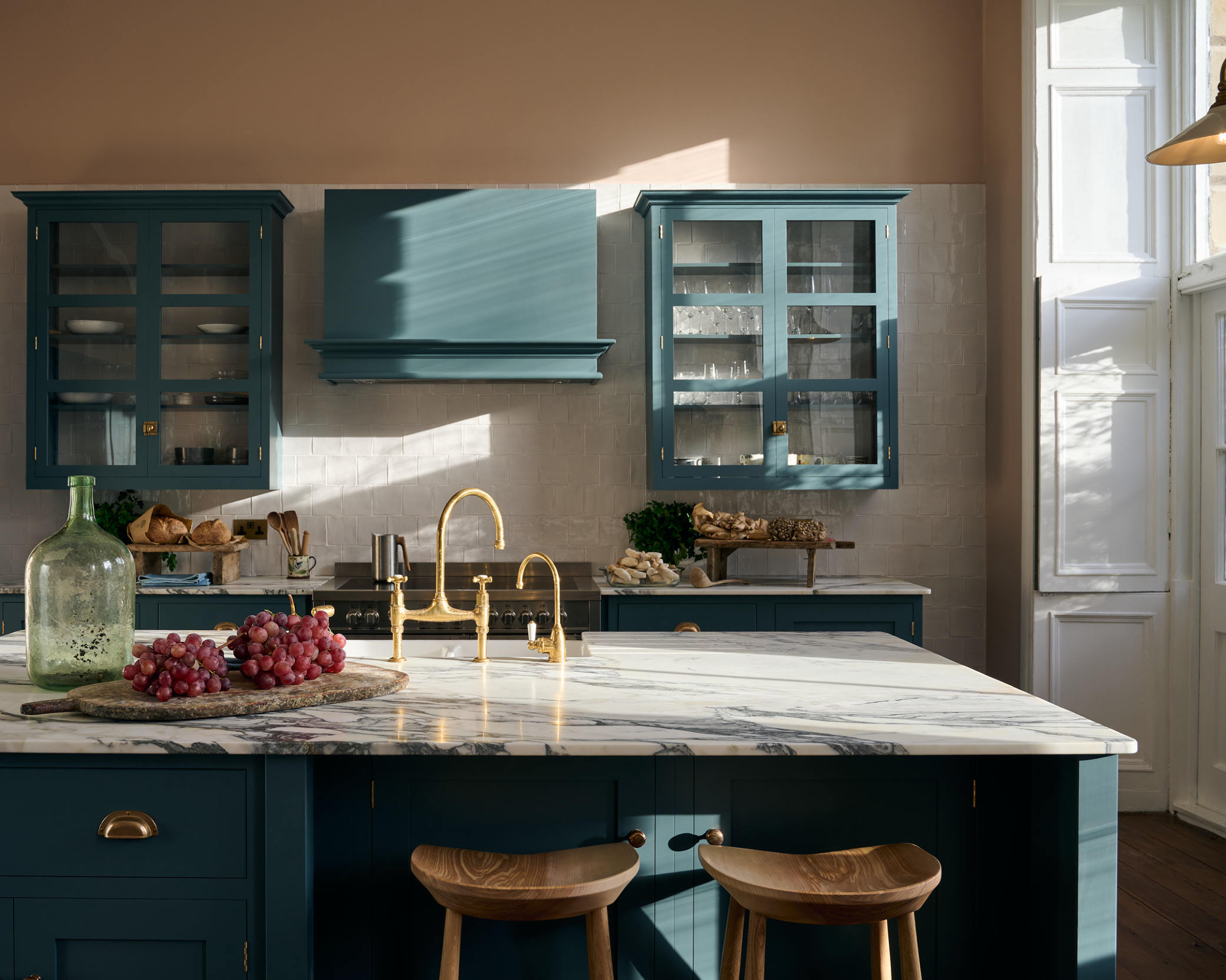 kitchen with teal cabinets, wall units with glazed doors, tiled backsplash, original window shutters, large kitchen island with marble countertop, bar stools, brass handles, painted cooker hood
