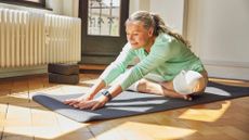 Woman learning how to loosen stiff joints, sitting on yoga mat at home in studio, sunshine coming through window