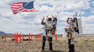 two people in skeleton-like spacesuits without the outer shell walk through the desert under a bright sun