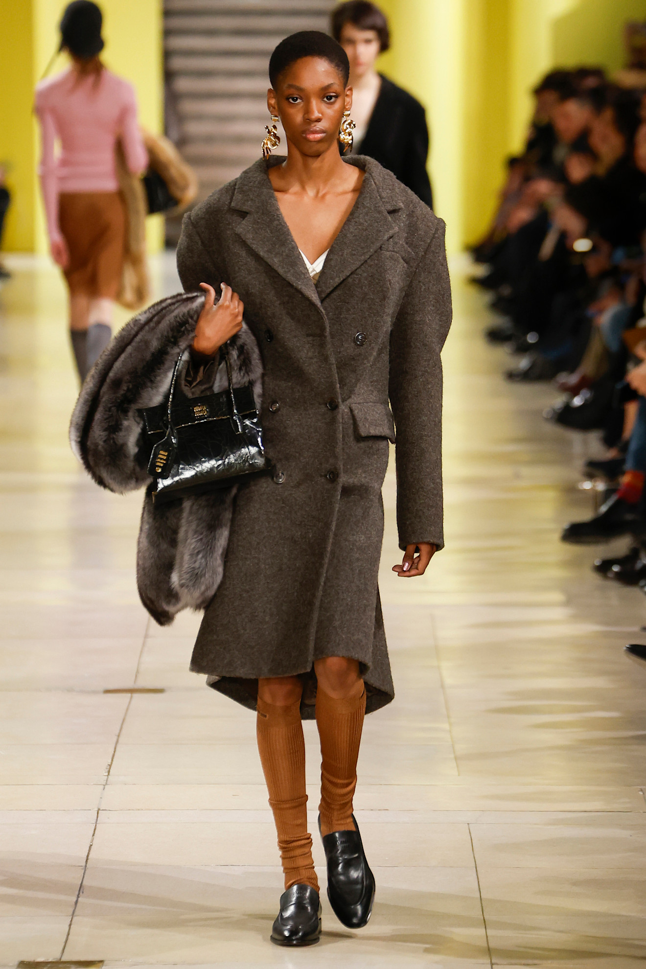 Fur accessory trend is shown in Miu Miu&#039;s fall-winter 2025 collection with a photo of a model wearing gold earrings with a white slip dress layered under an oversized brown wool coat styled with tan knee-high socks, black loafers, a black handbag, and a fur scarf