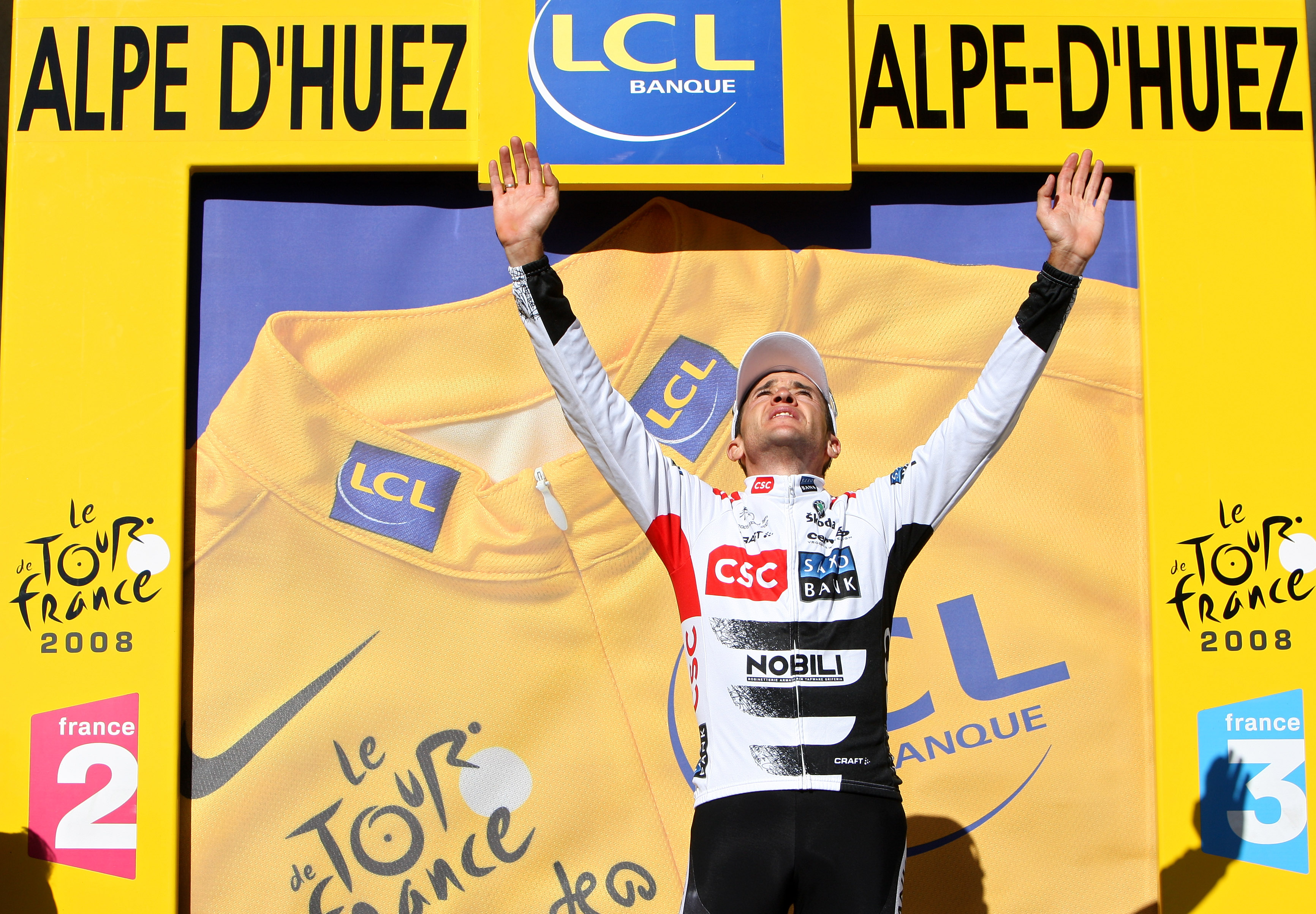 Carlos Sastre looks skyward with both arms raised on the Tour de France podium atop Alpe d'Huez in 2008