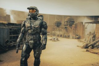 Master Chief (Pablo Schreiber) stands in a full suit of armour that makes him appear taller, stronger and more athletic than the average human. He is standing in the middle of an outpost run by a resistance group on a desert planet. There are several bodies lying on the ground behind him and damage to buildings that shows evidence of a recent battle.