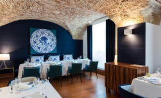 Inside look of the Ânfora restaurant. White walls and deep blue walls, with lowered exposed brick ceilings. Smaller tables against the back wall, covered in white linen and deep blue velvet chairs.