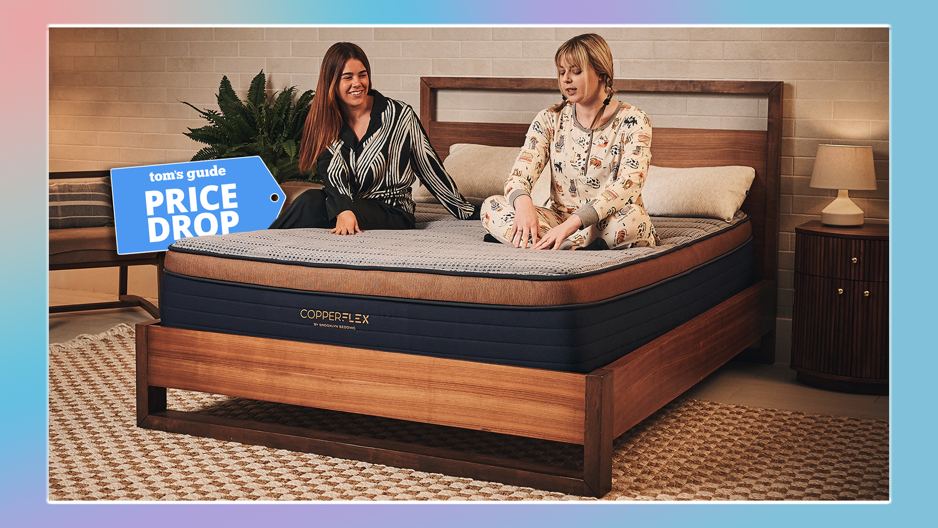 Two women sit on the Brooklyn Bedding CopperFlex mattress in a warmly lit room. Behind the mattress is a Tom's Guide Price Drop deals graphic. A colorful border surrounds the image