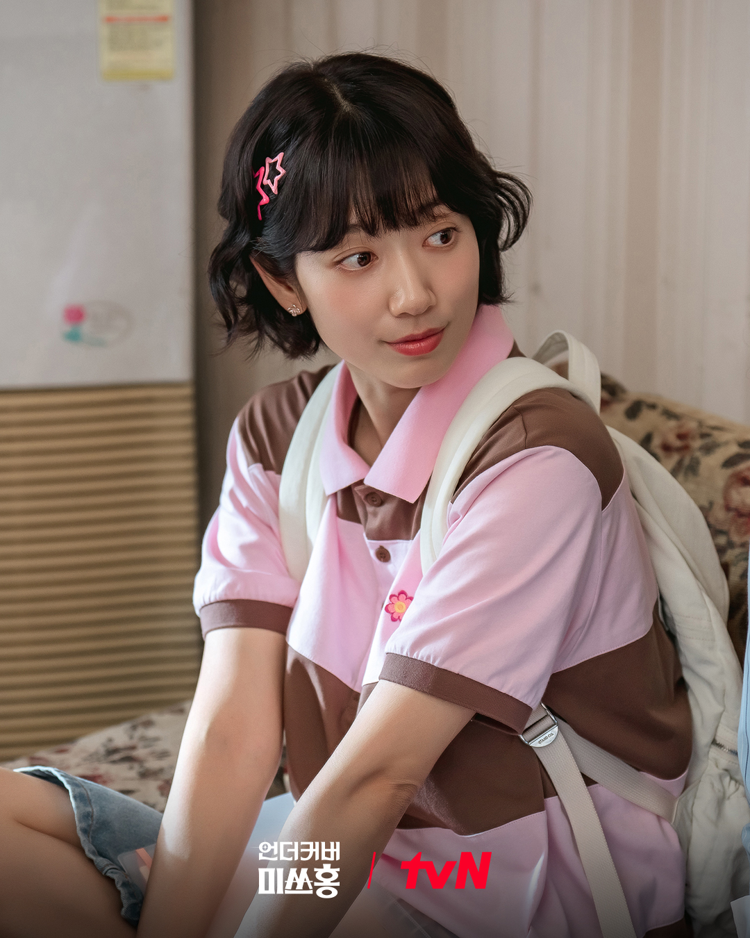 A thirty-something woman dressed like a '90s teenager (Park Shin-hye as Hong Keum-bo) sits on a floral couch, in the Netflix K-drama 'Undercover Miss Hong.'