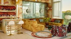 70s style living room with geometric wallpaper, mustard velvet sofa, colorful rug, patterned armchair, built-in home bar with bar stools, and shelves lined with liquors