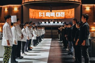 A row of White Spoon chefs face off against a row of Black Spoon chefs on a kitchen floor divided in half in white and black, with a round table and LED screen behind them, in 'Culinary Class Wars.'