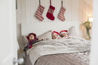 Three children in a double bed, with Christmas stockings hanging on the wall above
