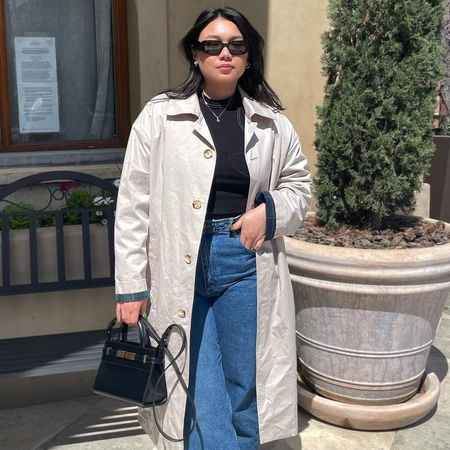 female mid size fashion influencer Marina Torres poses outside a building wearing black retro oval Prada sunglasses, a black mock neck top, light beige trench coat, high-waisted straight-leg jeans, and a mini black YSL bag