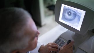 photo shows an eye doctor in the foreground, looking towards a computer monitor in the foreground that's showing a close up image of a patient's eyeball
