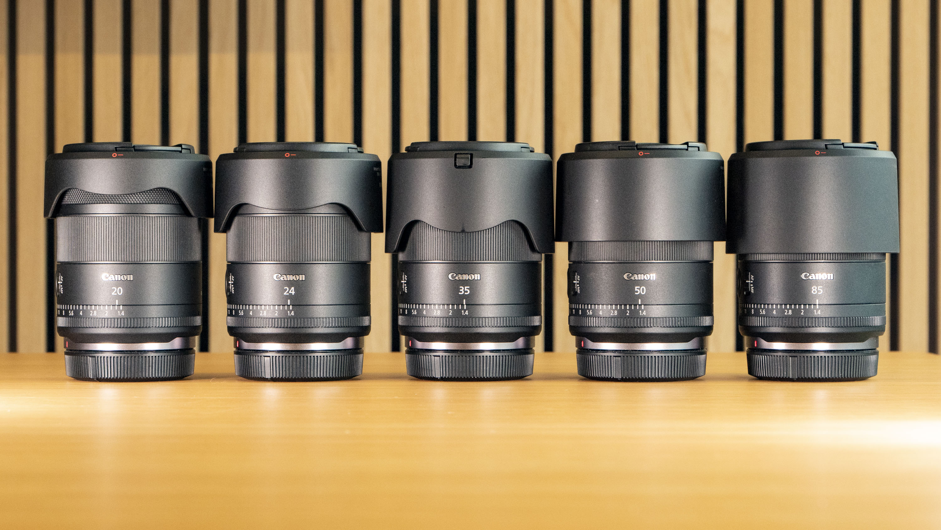 Canon RF 85mm F1.4L VCM lens on a wooden surface, alongside four other Canon F1.4 VCM lenses
