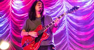 Mateus Asato plays a red Gretsch semi-hollow onstage in front of a pink-purple curtain.