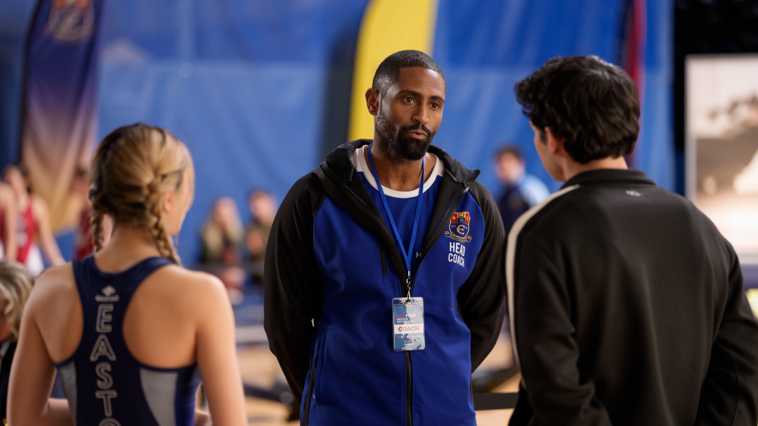 A high-school coach (Thomas Cadrot as Coach Hayden) stands in front of two teen athletes (Miku Martineau as Teagan and Kyle Clark as Cam Dillinger) in a gym, in Netflix's 'Crew Girl'.