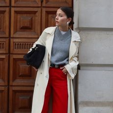 Influencer Alice Satterthwaite wears red jeans, a grey high-V-neck sweater, a long trench coat, pointed-toe boots, and a black suede tote. 