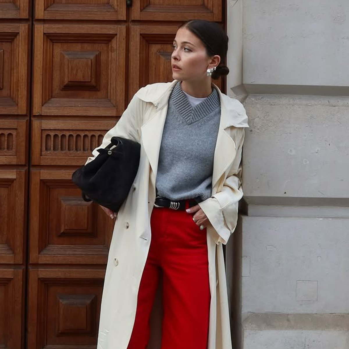 Influencer Alice Satterthwaite wears red jeans, a grey high-V-neck sweater, a long trench coat, pointed-toe boots, and a black suede tote. 