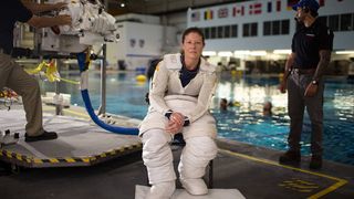 an astronaut in a spacesuit in front of a large pool. personnel stand in the background, in shadow