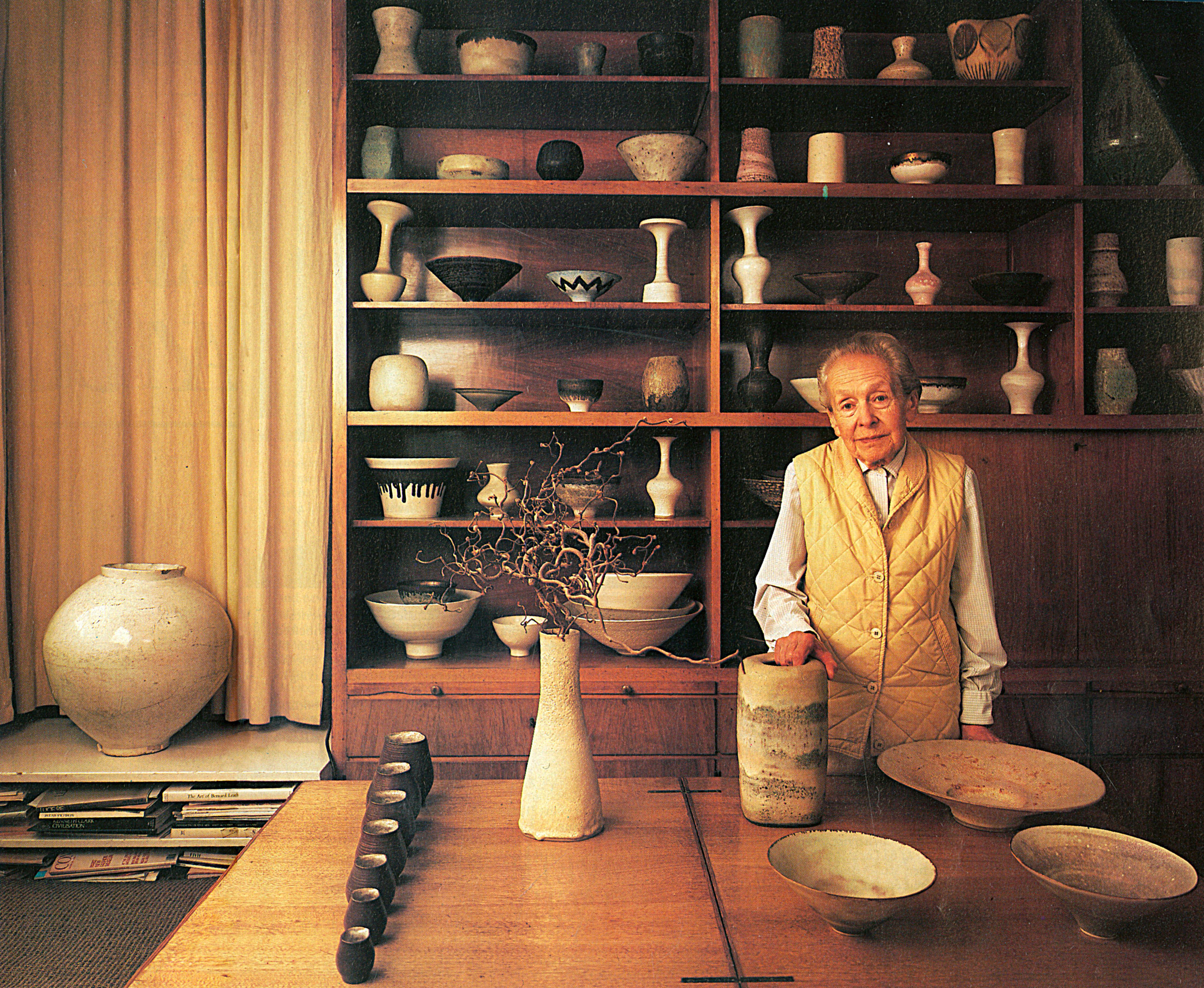 Lucy Rie wearing a yellow waistcoat and standing infront of a table of her famous bowls
