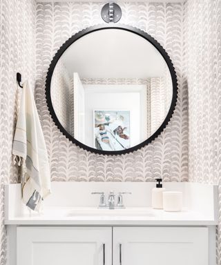 A white bathroom sink with a towel on the wall, white and gray wavy wallpaper, and a black circular mirror above the sink