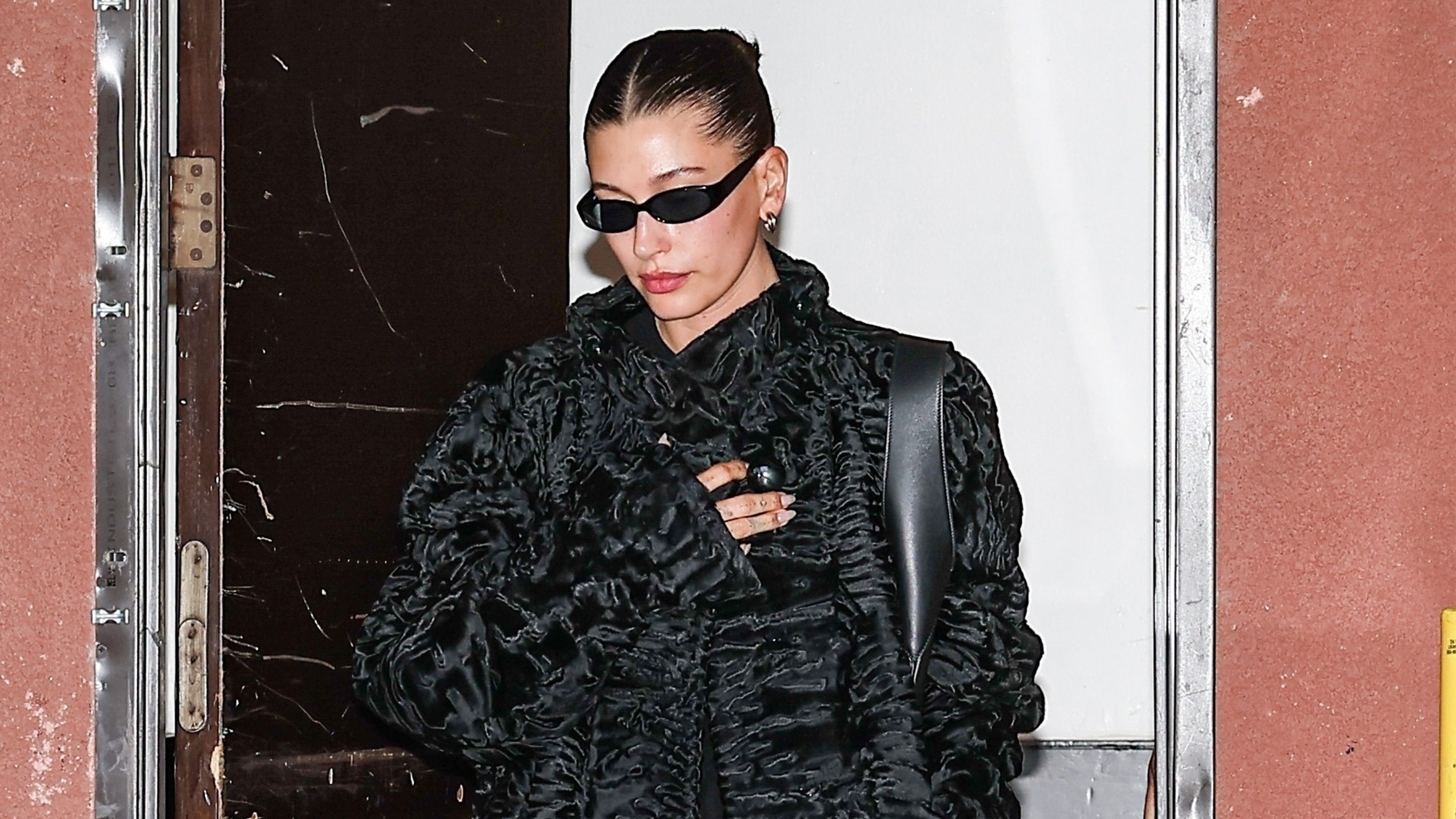 Hailey Bieber wears a black coat, a black leather bag, and black sunglasses.
