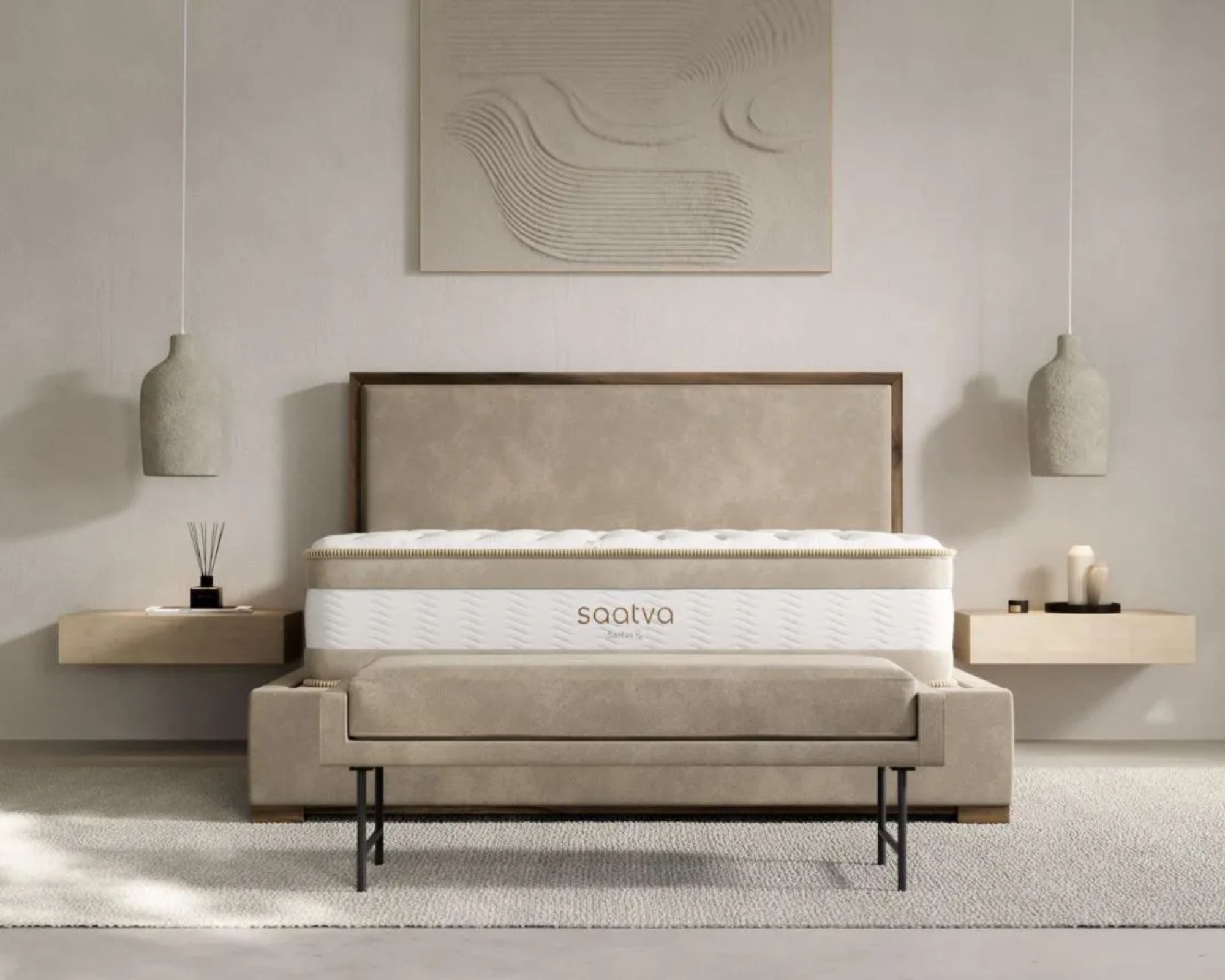 A Saatva RX mattress shown in a neutral bedroom with floating bedside shelves, a large artwork, hanging pendant lamps, and a beige bench with black legs.