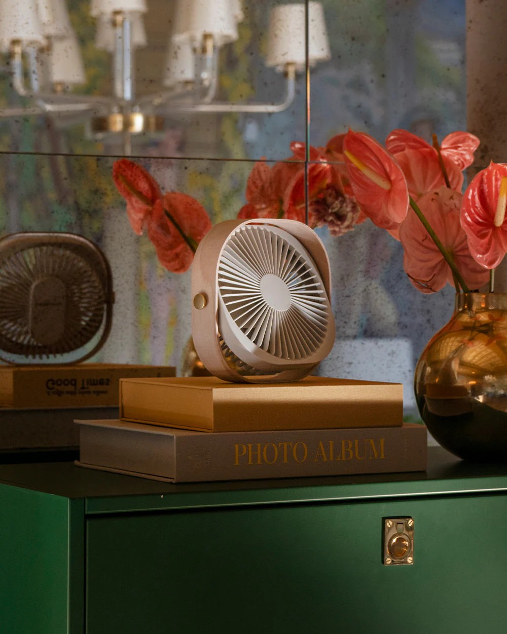 A small white and wooden fan sitting on a stack of coffee table books on a kelly green cabinet. The wall behind the cabinet is mirrored and there is a gold vase with orange flowers sitting beside the fan.