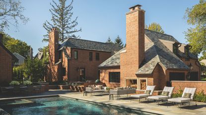 A luxury brick estate features a large swimming pool and a flagstone patio. The outdoor living area includes a massive brick fireplace with built-in wood storage, modern lounge chairs, and a sleek outdoor kitchen.