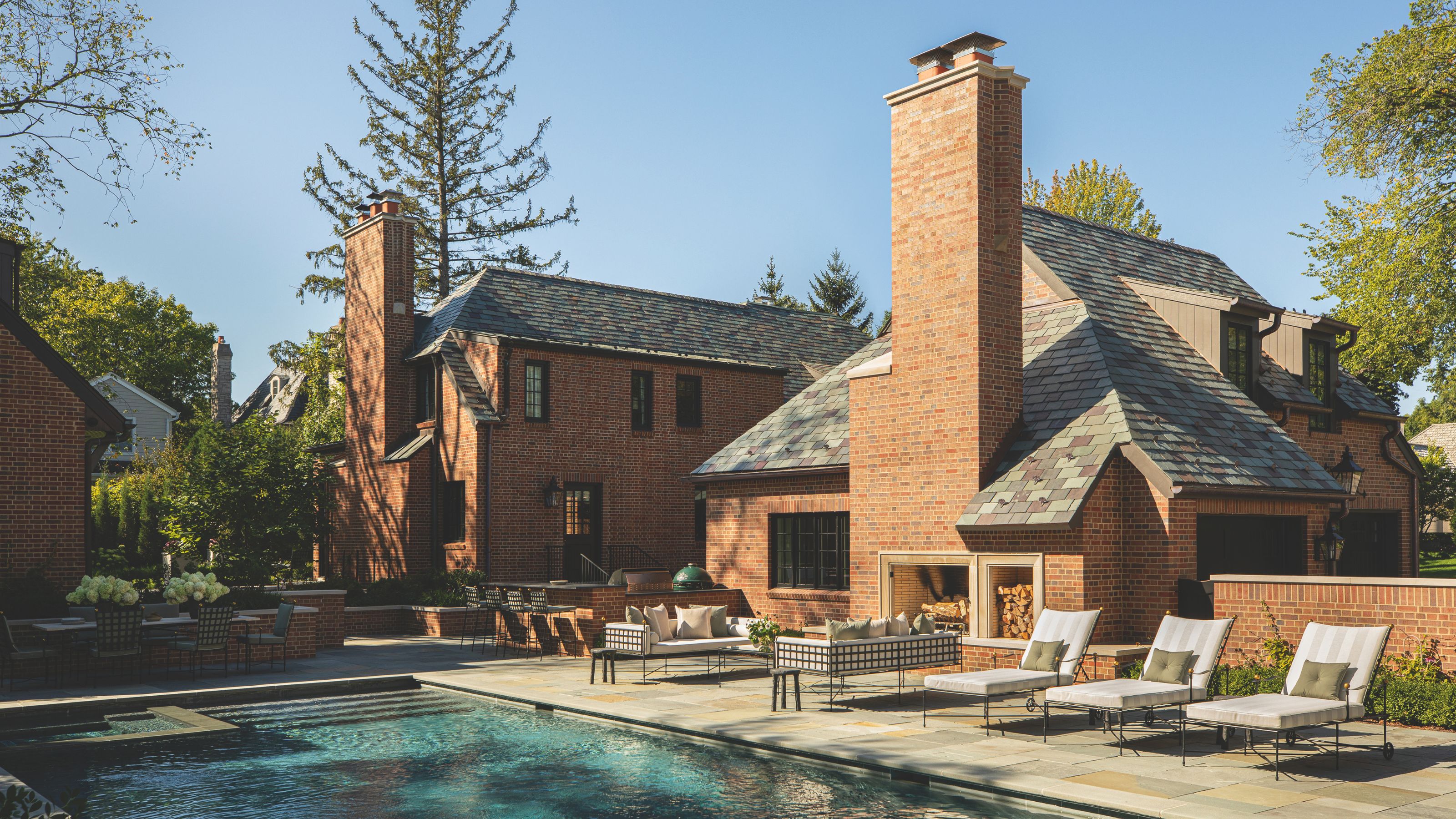 A luxury brick estate features a large swimming pool and a flagstone patio. The outdoor living area includes a massive brick fireplace with built-in wood storage, modern lounge chairs, and a sleek outdoor kitchen.