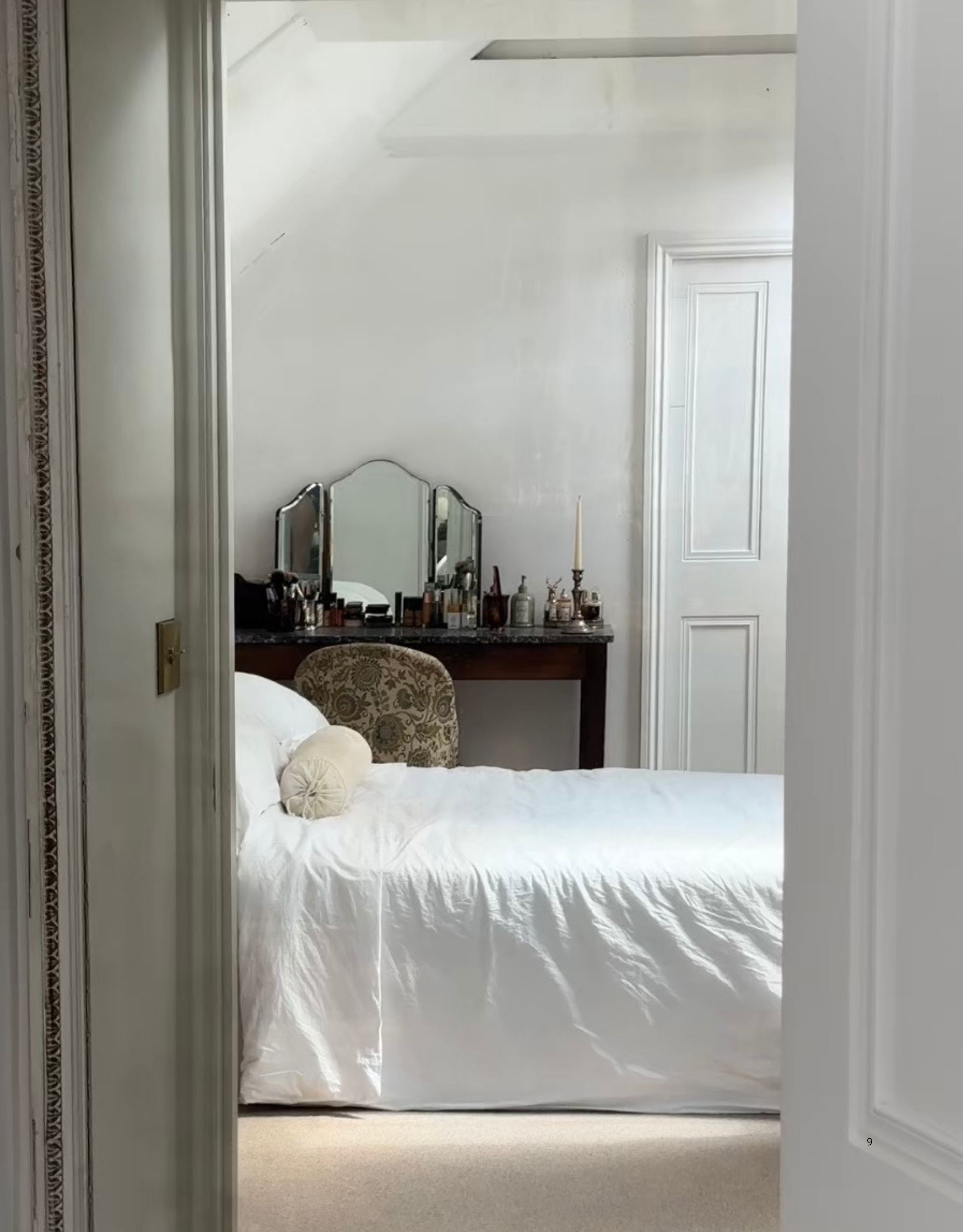 Image of a white bedroom with a white bed as seen through a doorway. There is a vanity at the back of the room full of different items, a mirror, and a patterned chair in front of it.