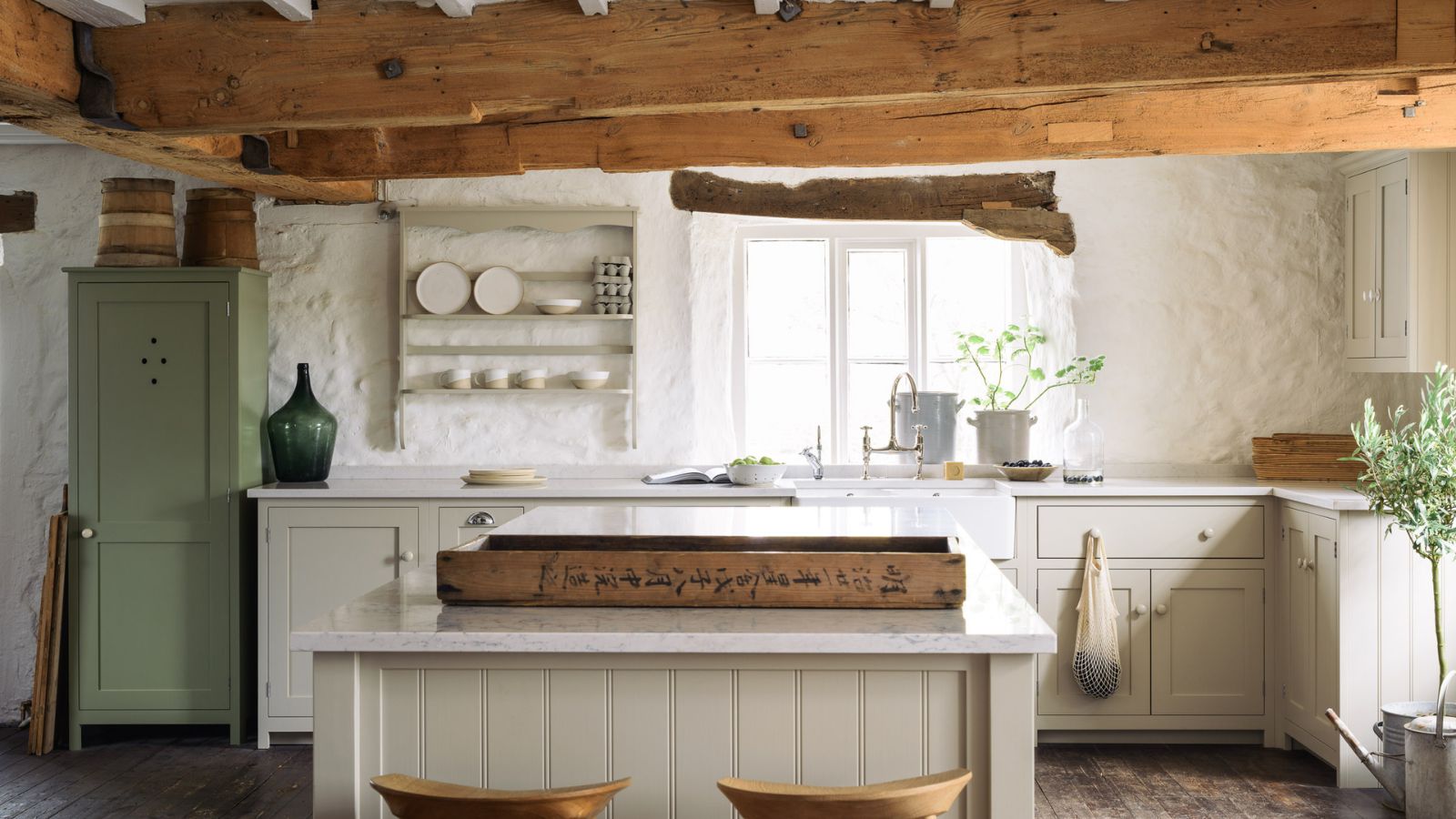 An English country kitchen with with white cabinets, a green freestanding cupboard, and exposed wood ceiling beams