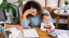 Stressed mother holds her baby at a desk with a calculator and laptop as she looks at credit card bills 