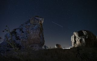 A long perseid meteor train streaks across the sky between two large rock structures in the foreground of the image.