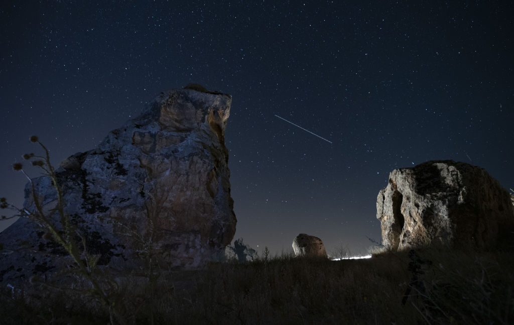 A long meteor train stretches across the sky between two large rock structures in the foreground of the image.