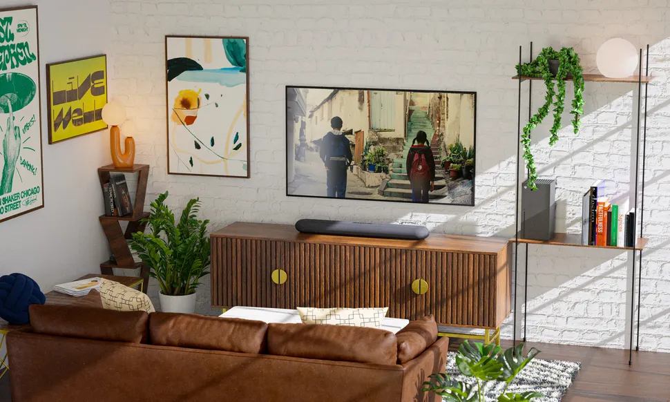 A lifestyle photo showing the Bluesound Pulse Cinema Mini soundbar on top of a TV unit beneath a wall-mounted TV. The room has white brick walls, dark furniture and several posters and pot plants