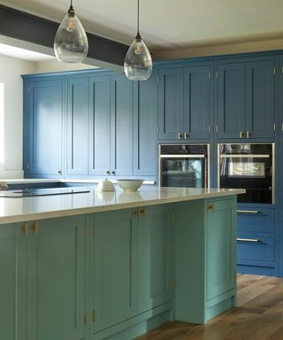 A light green kitchen island with a white countertop, two glass pendant lights above it, and a wall of blue cabinets with two black ovens